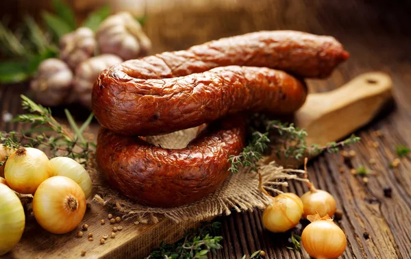 Salsiccia affumicata su un tavolo rustico in legno con aggiunta di erbe aromatiche fresche e spezie, prodotto naturale da agricoltura biologica, prodotto con metodi tradizionali — Foto Stock