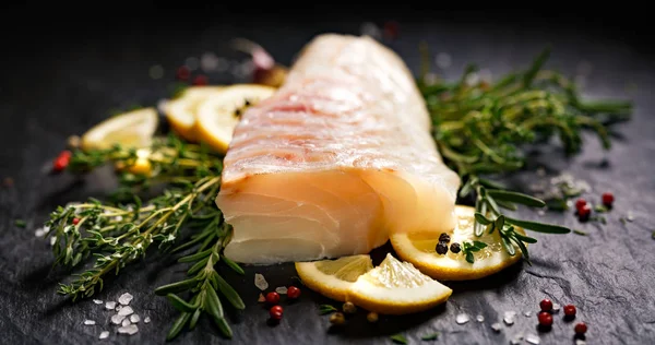 Fresh fish,  raw cod fillet with addition of herbs and lemon slices on black stone background, top view — Stock Photo, Image