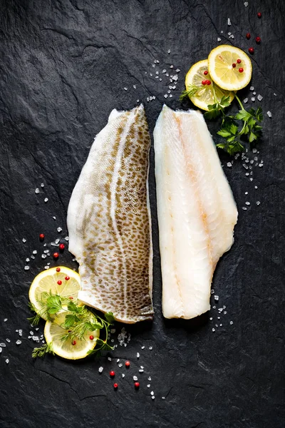 Peixe fresco, filé de bacalhau cru com adição de ervas e fatias de limão no fundo de pedra preta, vista superior — Fotografia de Stock