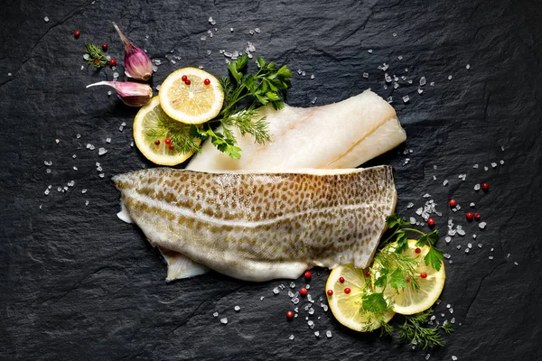 Friss hal, nyers tőkehal filé gyógynövények és citromos szelet a fekete kő háttér, felülnézet — Stock Fotó