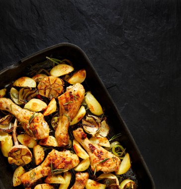 Patates dilimleri, sarımsak, soğan ve aromatik otlar bir ızgara tava, üstten görünüm, siyah taş zemin, kompozisyon köşedeki ızgara tavuk budu