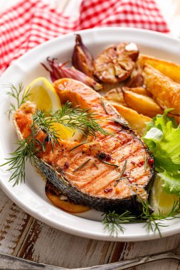 Grilled salmon steak, a portion of grilled salmon with fresh lettuce and potato wedges on a white ceramic plate on a wooden rustic table, close-up. Grilled seafood. clipart
