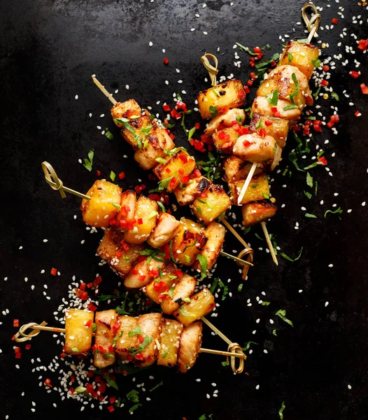 Grilled skewers with pineapple fruit and chicken meat  sprinkled with sesame seeds, chilli pepper and fresh herbs on a black background, top view . Fruit and meat skewers