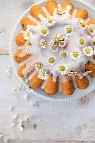 Easter yeast cake (Babka) covered with icing and decorated with daisies on a white plate on a white wooden table, top view. Traditional Easter cake in Poland