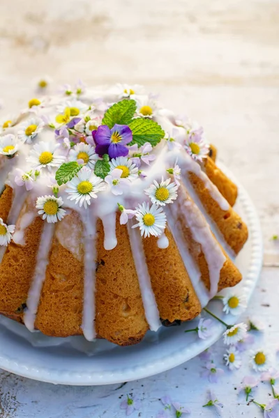 Easter yeast cake (Babka) covered with icing and decorated with edible flowers on a white plate on a white wooden table, close up. Traditional Easter cake in Poland