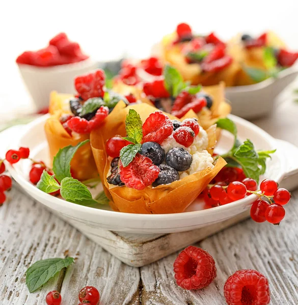 Phyllo pastry fruit cups with whipped cream filling topped with mix fresh berries sprinkled with powder sugar on a white plate close up. Delicious filo pastry dessert