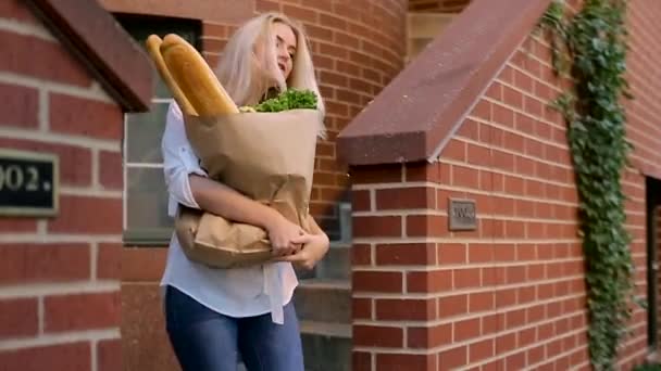 Junges blondes Mädchen kommt die Treppe hinunter und ist in den Händen einer Papiertüte mit Lebensmitteln, die Brot und Kräuter ist, riecht und lächelt. — Stockvideo
