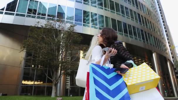 Duas meninas de pé na rua perto do centro comercial com compras, segure o telefone, abraçando, beijando e rindo . — Vídeo de Stock