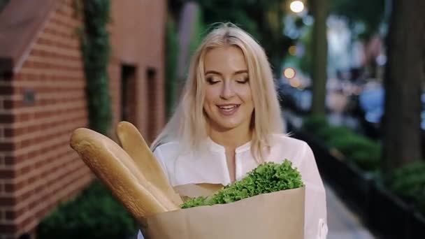 Молода блондинка стоїть на тротуарі і тримає пакет продуктів . — стокове відео