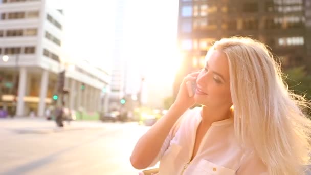 Νεαρή ξανθιά κοπέλα στέκεται κοντά σε μεγάλα κτίρια όπου ο ήλιος λάμπει φωτεινός. Slow-Motion. Μιλάει στο τηλέφωνο. — Αρχείο Βίντεο