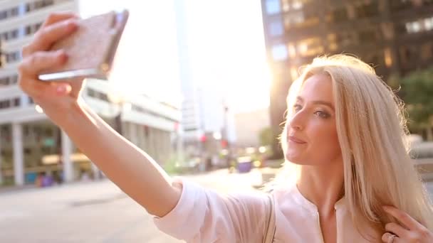 Una joven rubia haciendo selfie, está en el camino cerca de edificios altos . — Vídeos de Stock