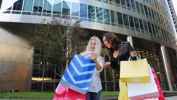 Twee jonge vrouwen permanent met allerlei pakketten rond het gebouw en een enorme lach. Communiceren en vreugdevol kijken naar de telefoon. — Stockvideo