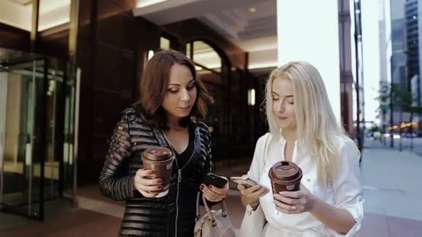 Twee jonge meisjes permanent in de buurt van een groot gebouw en houden kopjes koffie en telefoons. — Stockvideo