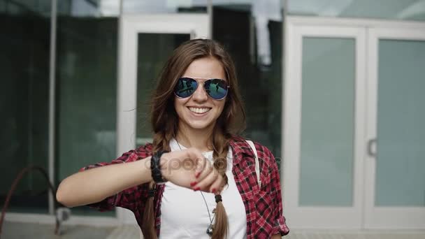 Joyeuse jeune femme aux cheveux tressés bruns et des lunettes modernes et des vêtements décontractés en regardant sa montre fraîche près de l'entrée du bâtiment — Video