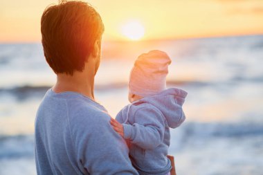 Kollarında bebek olan bir adam arkasında duruyor ve gün batımını seyrediyor.