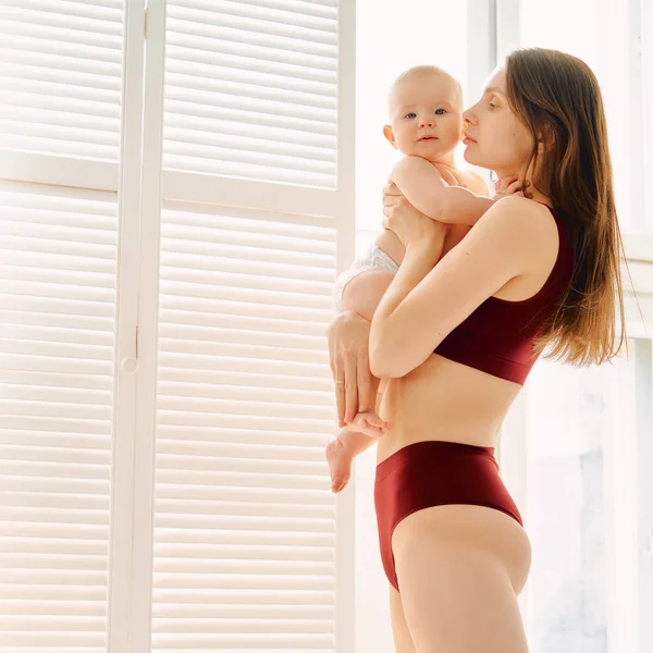 Mãe com um bebê em seus braços olha para ele no fundo da janela — Fotografia de Stock