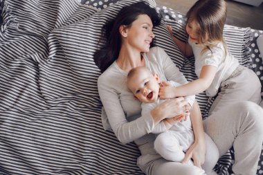 Genç, gülümseyen bir anne çizgili bir yatakta iki küçük kızla oynar. Çocuklar gülüyor.