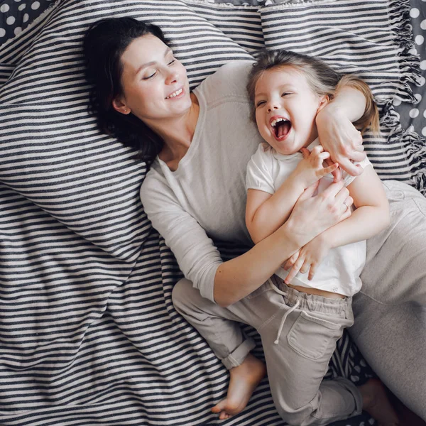 Bela mãe com 3 anos de idade filha abraçar e rir enquanto deitado na cama listrada — Fotografia de Stock