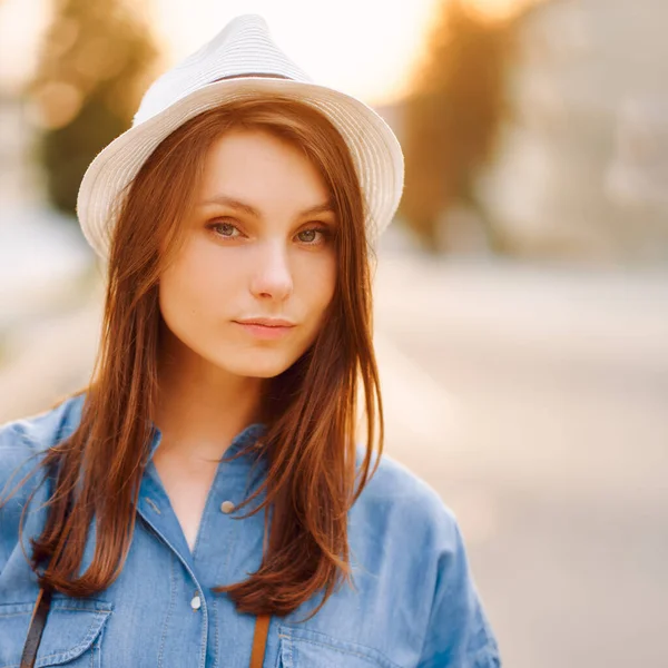 Portret dziewczyny w białym kapeluszu i niebieskiej koszuli jeansowej na tle miasta zachodu słońca — Zdjęcie stockowe