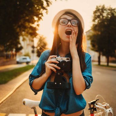 Şapkalı şık bir kadın sabah güneşinde fotoğraf makinesiyle bisikletin yanında duruyor.
