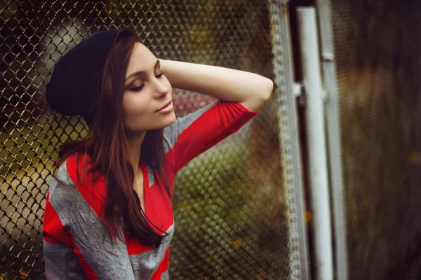 Portrait of a beautiful girl in a hipster hat and striped jacket on the street at the gate outdoor — Stock Photo, Image