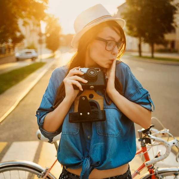 Stylowa kobieta w kapeluszu stojąca w pobliżu roweru w porannym słońcu z aparatem fotograficznym na zewnątrz — Zdjęcie stockowe