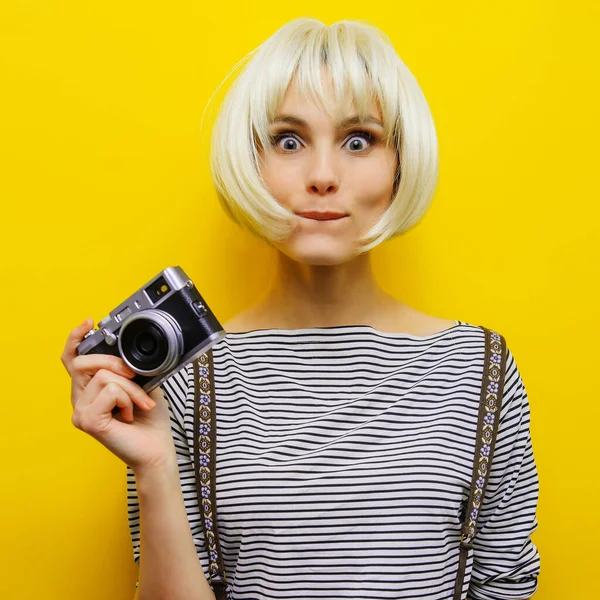 Portret uśmiechniętej dziewczyny z kamerą w dłoni na żółtym tle. Odizolowane studio. peruka blond — Zdjęcie stockowe