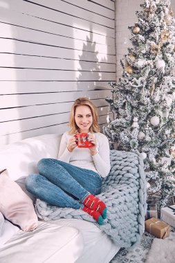 Güzel sarışın bir kadın, dekore edilmiş bir yılbaşı ağacının arka planında kırmızı bir fincanla bir kanepede oturuyor. Kız Noel tatilinde dinleniyor.