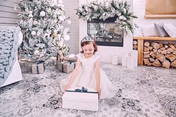 Beyaz elbiseli küçük bir kız elinde hediyesiyle bir Noel ağacının arka planında İskandinav iç mimarisi olan bir odada bir kutu tutuyor. Yılbaşı tatilinde çocukların neşesi.