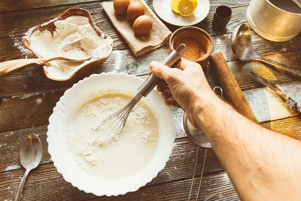 Der Koch knetet den Teig mit einem Schneebesen. Weizenmehl, Teig, Eier, eine Zitrone und Küchengeräte auf einem Holztisch. Zubereitung des Teiges in einer rustikalen Küche. getöntes Bild — Stockfoto