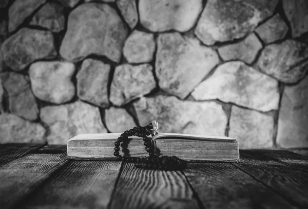 Różaniec z krzyżem na otwartej księdze starego na starym drewnianym stole na tle ściany z kamienia. Selektywny fokus, czarna idealna białe zdjęcie. z miejsca na tekst — Zdjęcie stockowe