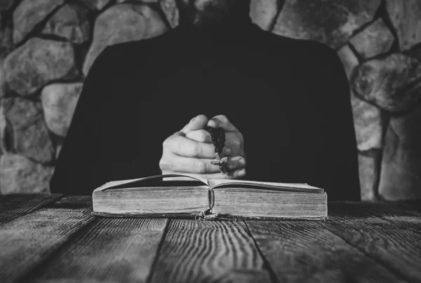 Um homem de roupas pretas com contas de oração na mão rezando na frente de um velho livro aberto. o conceito de orar e estudar. foco seletivo, foto preto e branco — Fotografia de Stock