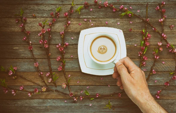 Cup with coffee milk in a male hand on a background of an old wooden table with peach branches with pink flowers, top view. Retro toning photo with vignette — Stock Photo, Image
