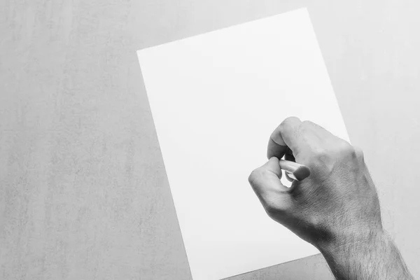 Mão macho com uma caneta esferográfica e uma folha de papel branca em branco sobre fundo cinza, vista superior close-up. foto em preto e branco. mock up para texto, parabéns, frases, letras — Fotografia de Stock