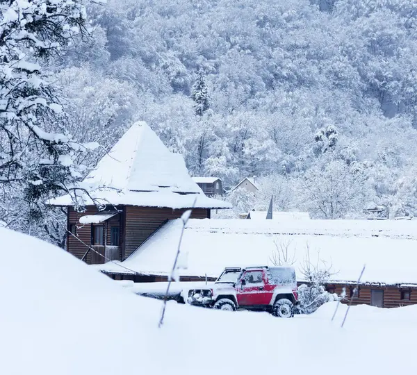 Winterlandschap met een auto is een rode SUV in de sneeuw in de buurt van een houten huis op een achtergrond van bos heuvels in de wintertijd vroeg in de ochtend — Stockfoto