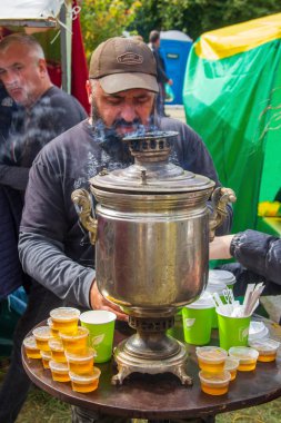 Adigea, Rusya - 21 Eylül 2019: Sakallı bir adam semaver ve bal kavanozlarıyla bir masada duruyor ve Adighe peynir festivalinde turistler için çay hazırlıyor