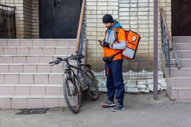 Krasnodar, Russia - February 11, 2020: young man courier service food delivery broniboy with the bike with the phone in his hand stands at the entrance of the house