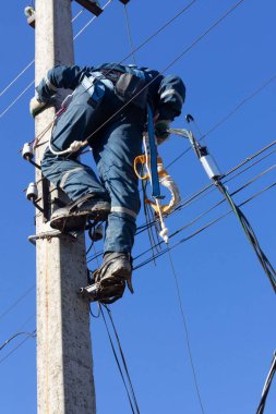 Erkek bir elektrikçi, elektrik kablolarından bir hat yapmak için güçlendirilmiş beton bir sütuna tırmanır.