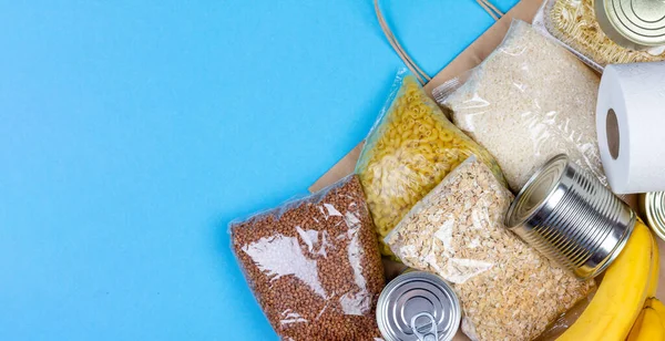 Paper bag with a crisis food supply for the period of quarantine isolation of the coronavirus, rice, pasta, oatmeal, canned food, toilet paper, buckwheat, bananas on a blue background with copy space