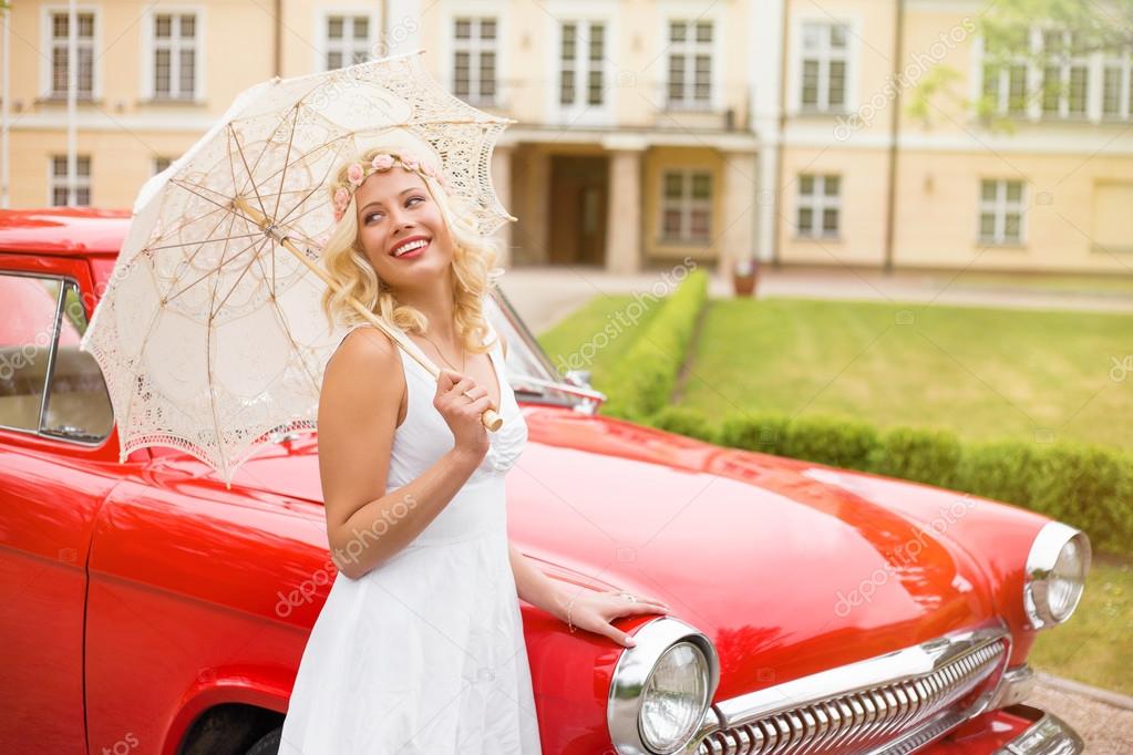 Woman in white dress standing next to red retro car