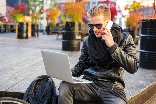 Hipster homem na cidade trabalhando no laptop e falando ao telefone — Fotografia de Stock