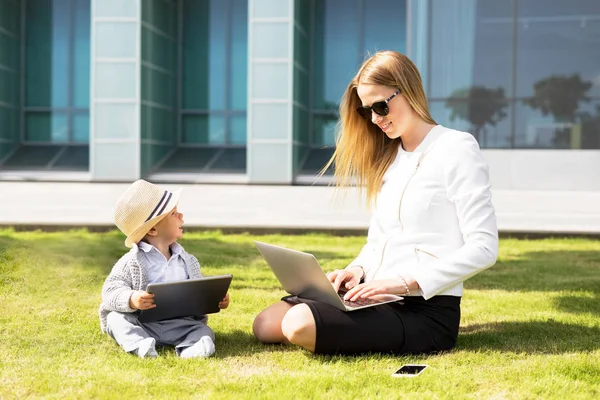 Ділова жінка зі своєю дитиною сидить на траві і працює над портативними інформаційними пристроями — стокове фото