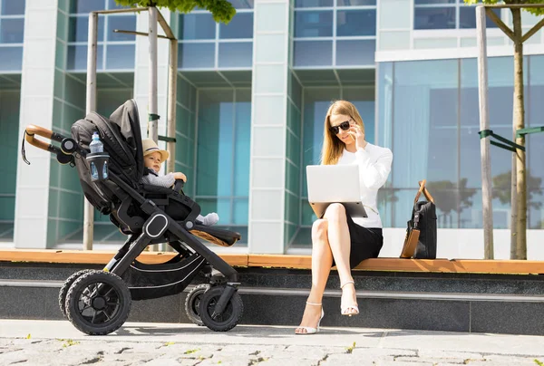 Business woman talking on the phone and working on laptop outdoors while having a baby in stroller — Stock Photo, Image