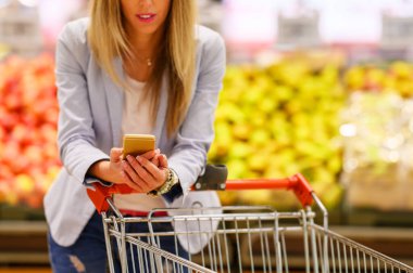 Kadın markette telefonuyla alışveriş listesini karıştırıyor.