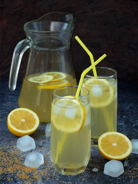 Zwei Gläser und eine Karaffe kalte hausgemachte Limonade mit Zitronenscheiben, Eiswürfeln, braunem Zucker, gelben Strohhalmen auf dunklem Hintergrund. — Stockfoto