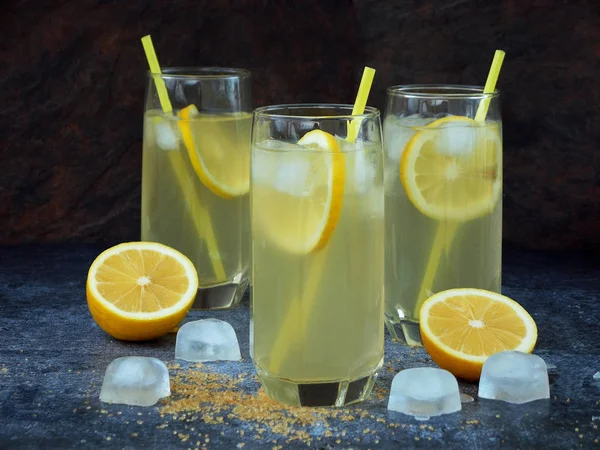 Drie glazen van koude zelfgemaakte limonade met citroen segmenten, ijsblokjes, bruine suiker en rietjes op donkere achtergrond. — Stockfoto