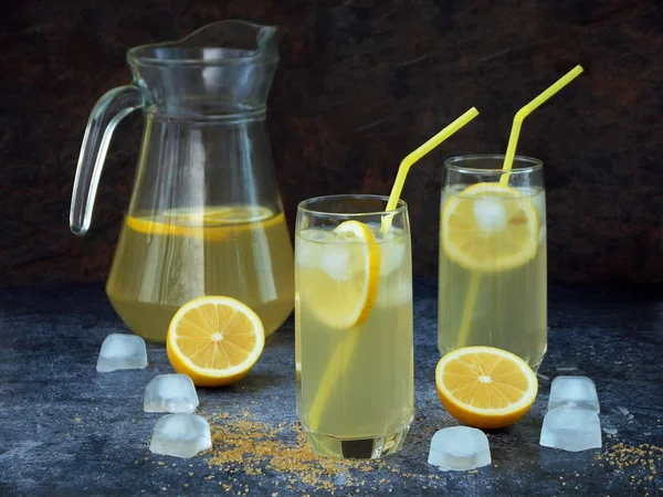 Два стакана и графин холодного домашнего лимонада с ломтиками лимона, кубиками льда, коричневым сахаром, желтыми соломинками на темном фоне . — стоковое фото