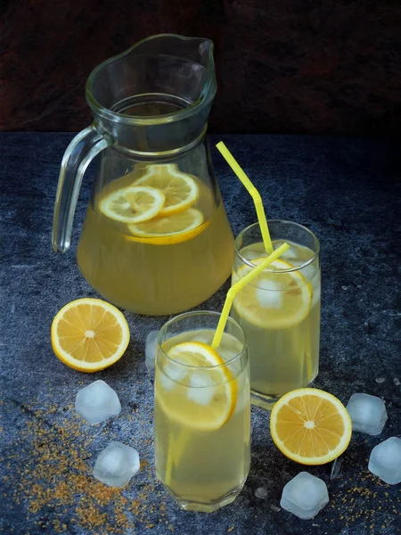 Два стакана и кувшин холодного домашнего лимонада с ломтиками лимона, кубиками льда, коричневым сахаром, желтыми соломинками на темном фоне . — стоковое фото