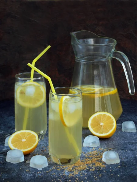 Zwei Gläser und ein Krug kalte hausgemachte Limonade mit Zitronenscheiben, Eiswürfeln, braunem Zucker, gelben Strohhalmen auf dunklem Hintergrund. — Stockfoto