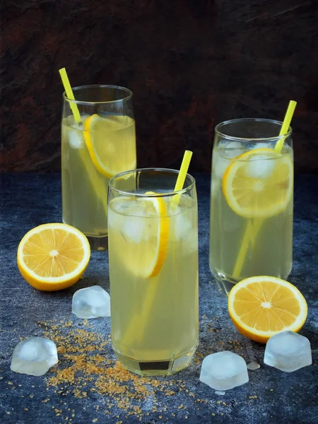 Drie glazen van koude zelfgemaakte limonade met citroen segmenten, ijsblokjes, bruine suiker en rietjes op donkere achtergrond. Kopiëren van ruimte — Stockfoto